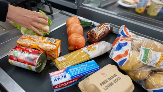 Stock image shows a range of items from a grocery shop on a supermarket conveyor belt, including eggs, potatoes, tipped tomatoes, and toothpaste