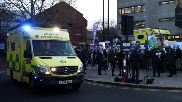 An ambulance with flashing blue lights drives past striking workers holding placards