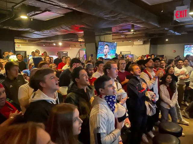 People standing watching TV screens in a crowded DC bar