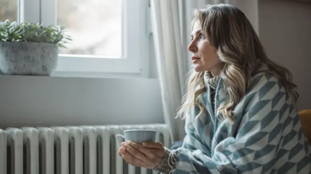 Woman in blanket siting sitting near a radiator and holding a mug