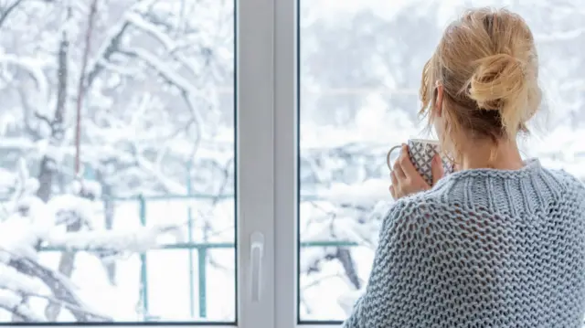 A woman stands at a window with a cup of drink in her hands as she looks out on snow