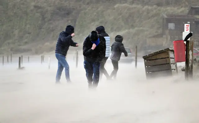 A group of people on the beach with hats and hoods pulled down over their faces to shield from the wind