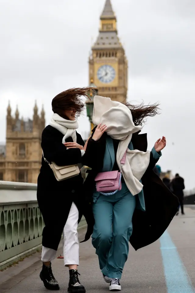 The wind whips a scarf into a woman's face as she and her friend cross Westminster Bridge, with Big Ben in the background