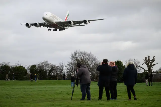 A group of four people in a field watch an Emirates plane coming overhead