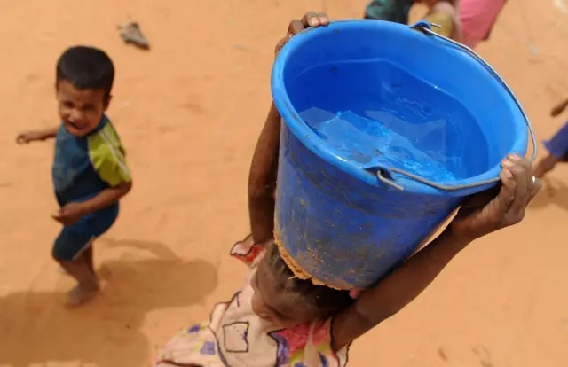 Children in Mauritania carry buckets of water as they walk across sand.