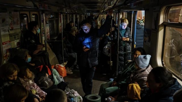 Man on dark train carriage using his phone surrounded by other people seeking shelter