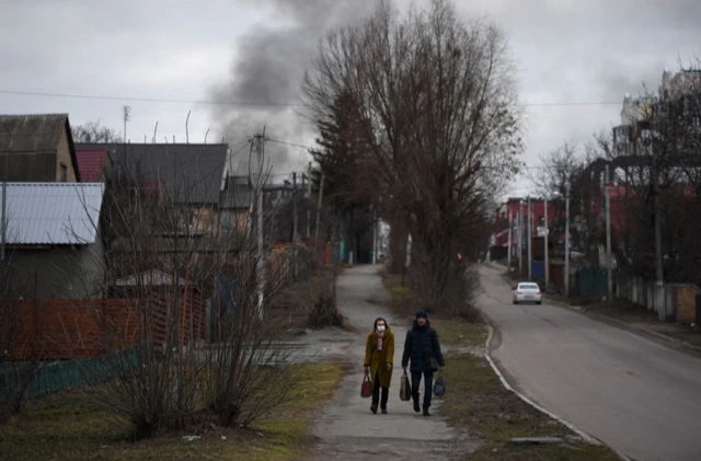 Local residents walk down a street as smoke rises near the Hostomel airfield