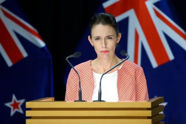 New Zealand Prime Minister Jacinda Ardern stands at lectern with backdrop of New Zealand flag