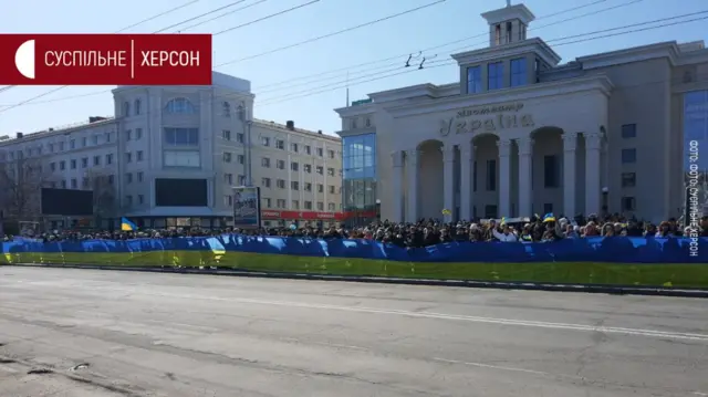 A line of people at a protest is seen holding a long Ukrainian flag