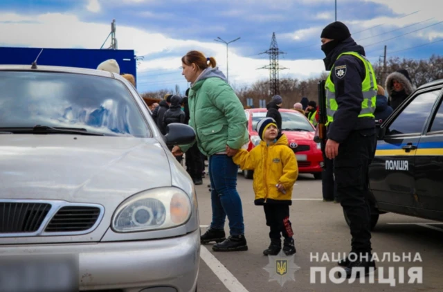 A woman holds a young boy's hand as they stand next to a police officer while in a refugee queue