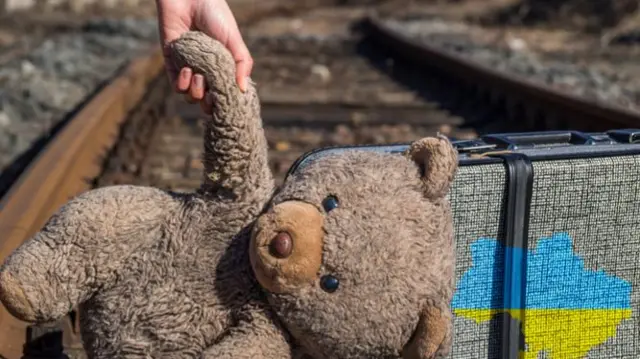 A child carrying a teddy bear next to a suitcase with a picture of a map of Ukraine on it.