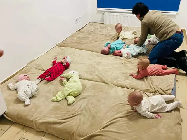 A group of infants lie on mattresses pushed together as a volunteer attends to one of them