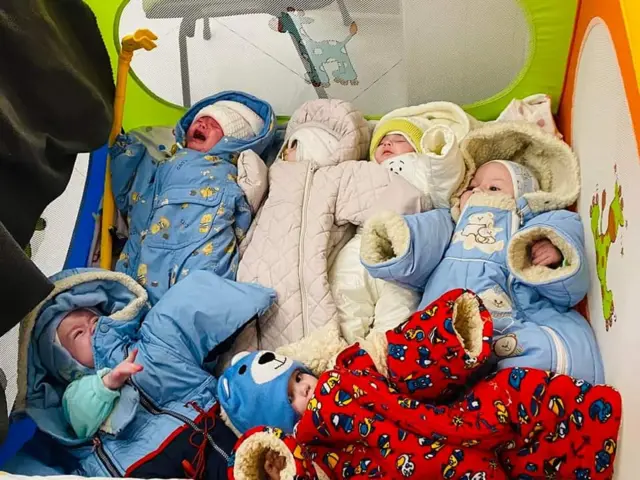 Six babes, bundled up in warm clothes, lie together in a cot