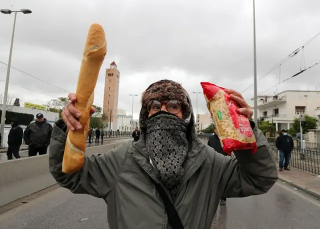 A Tunisian woman holds a baguette and pasta during a protest against Tunisian President Kais Saied"s seizure of governing powers, during the 66th anniversary of Tunisia"s independence in Tunis, Tunisia, 20 March 2022