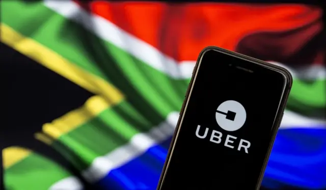 Uber application logo is seen on a screen in front of a flag of South Africa