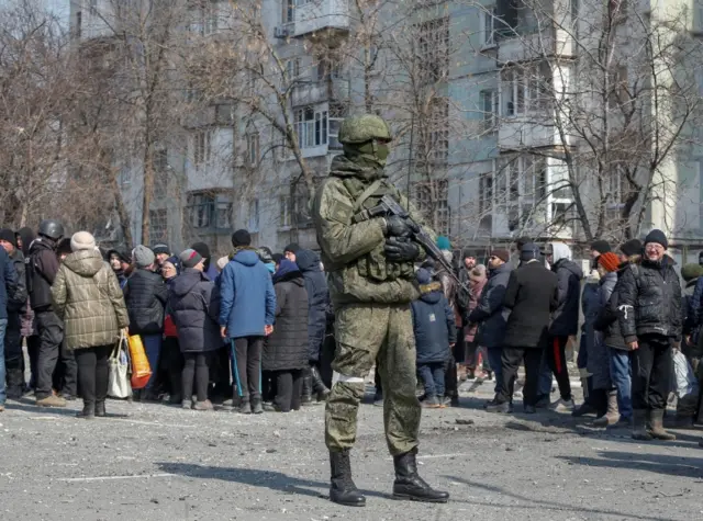 A Russian soldier stands guard as Mariupol residents who queue for humanitarian aid delivered by lorry