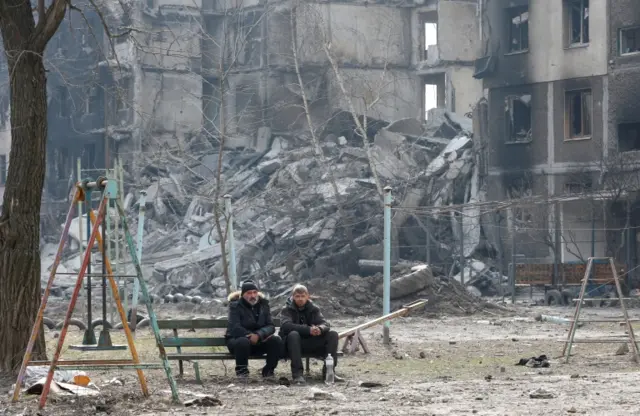 Local residents sit on a bench near a destroyed apartment building in the besieged city of Mariupol