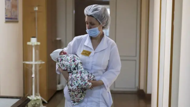 A nurse carries a baby down the corridor of a maternity hospital