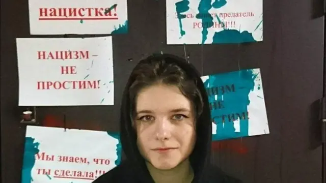 A young woman, Darya Kheikinen, in front of the vandalised door to her apartment