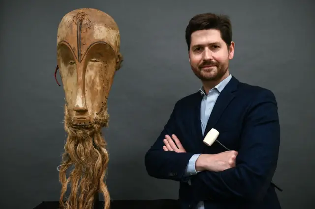 Auctioneer Jean-Christophe Giuseppi poses next to a "Ngil" mask of the Fang people of Gabon