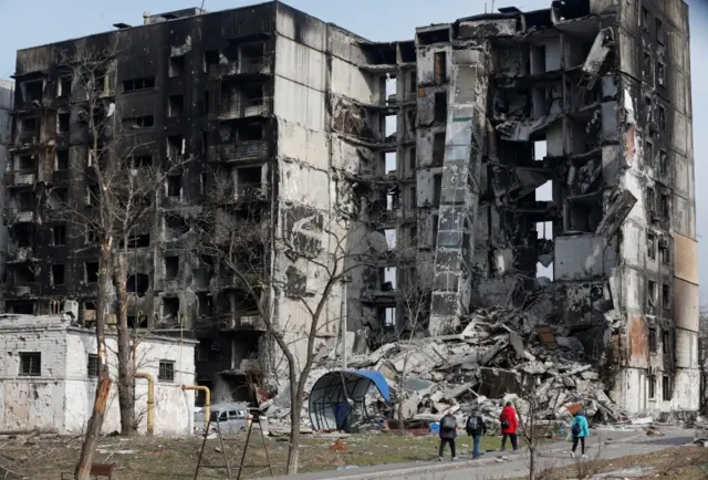 People walking by destroyed building in Mariupol