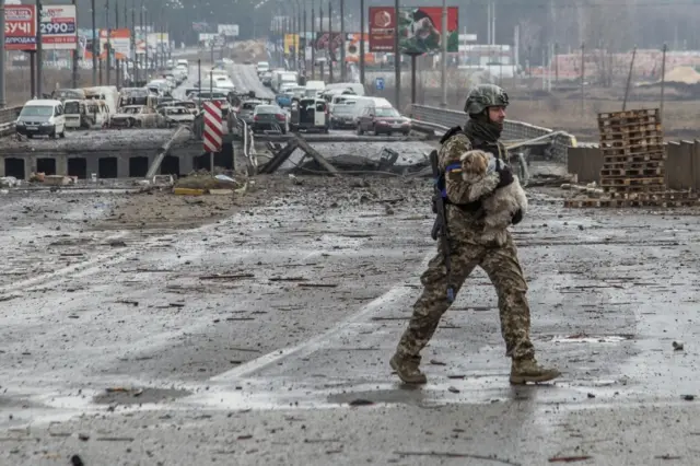 A Ukrainian service member carries a dog next to a destroyed bridge over the Irpin river