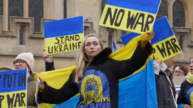 Olena in a 'I stand with Ukraine' hoodie, holding a Ukrainian flag and a placard saying "no war"