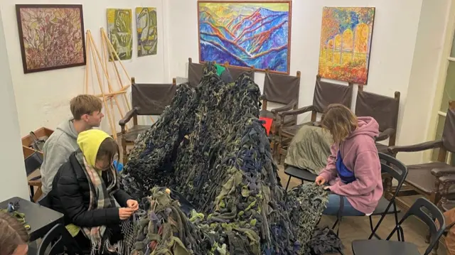 Three young people sit around a huge camouflage net, weaving more material into it in an art gallery