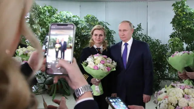 People take a photo of Putin stood next to a woman holding flowers