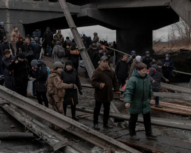 Ukrainians in military fatigues direct civilians over the remnants of a bridge