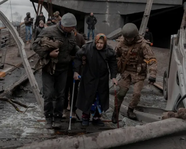 An elderly woman is helped while crossing a destroyed bridge as she tries to leave the city of Irpin