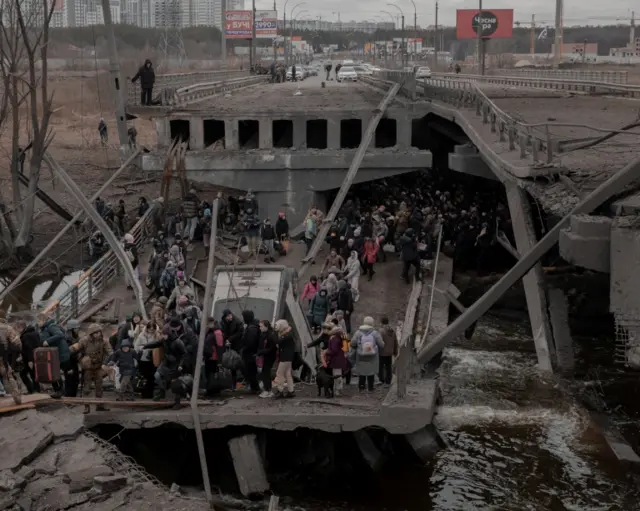 The river can be seen as people wait their turn to cross the destroyed concrete bridge