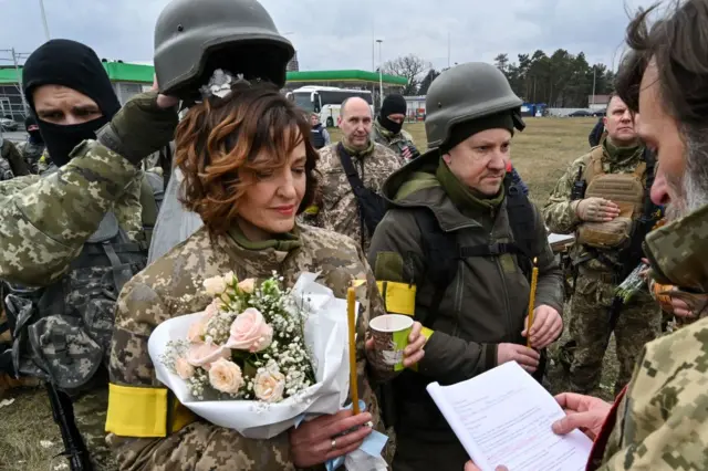 Lesya and Valery stand alongside their fellow soldiers, carrying candles and flowers, as a soldier holds Lesya's helmet over her head to protect her wedding veil