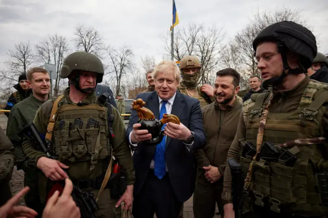 Boris Johnson holds a ceramic cockerel while walking with Volodymyr Zelensky and Ukrainian soldiers