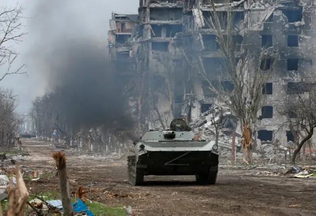 Russian troops ride an armoured vehicle through a decimated street in Mariupol