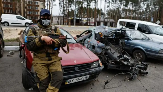 A member of the Ukrainian Territorial Defence Forces sits next to a destroyed car in Irpin