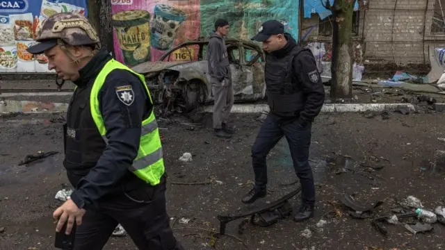 Police officers and a man walk past a burned car in the residential area that was hit by the Russian artillery shelling, in Kharkiv