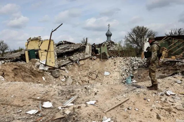 A Ukranian serviceman looks into a crater and a destroyed home are pictured in the village of Yatskivka, eastern Ukraine