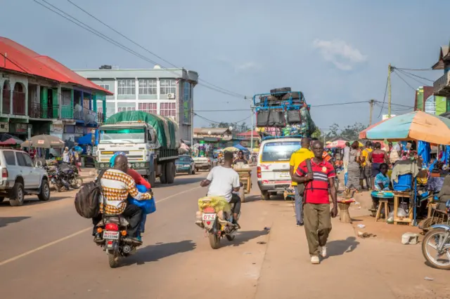 Cars, motorbikes, minibuses and lorries drive next to roadside vendors in Ganta, Liberia, in 2018.