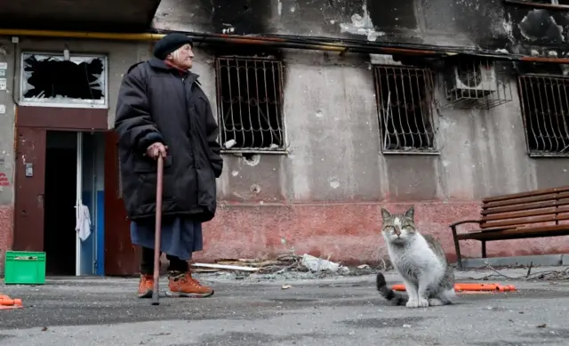 Maria Miroshnichenko, 84, a pensioner and former social service worker, stands near a cat outside a residential building heavily damaged during Ukraine-Russia conflict in the southern port city of Mariupol on 22 April 2022