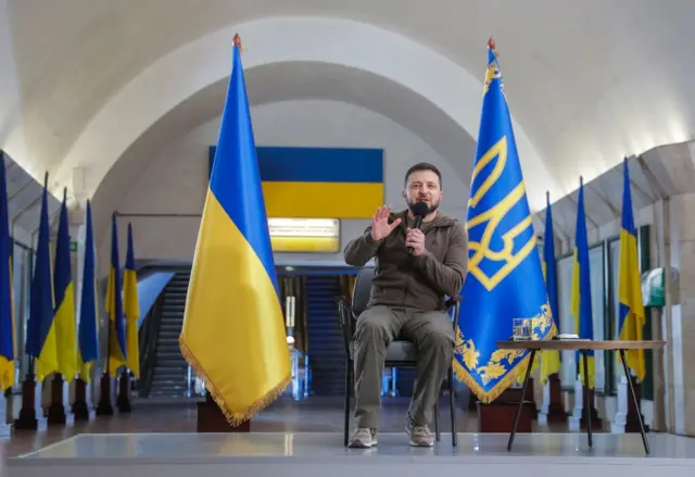 President Zelensky speaks to reporters in front of Ukrainian flags in a metro station