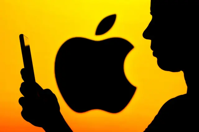 In this photo illustration, the Apple Inc logo is seen in the background of a woman's silhouette holding a mobile phone