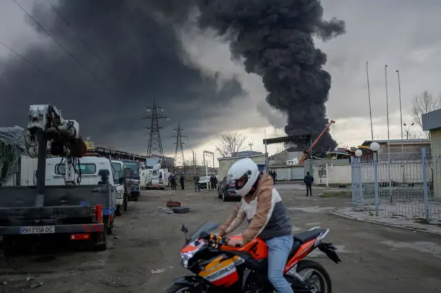 Image shows man on motorcycle in front of thick smoke rising out of ground in distance