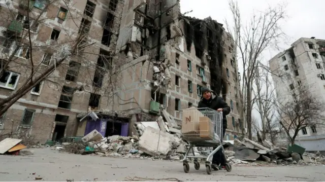 A resident pushes a trolley past a damaged apartment building in Mariupol