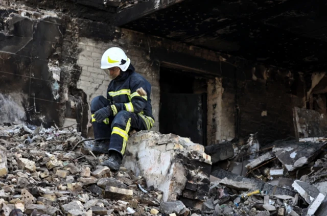 A rescuer rests on a pile of rubble outside a bombed building