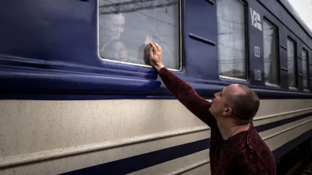 A man holds his hand up to a train window with a woman and child crying inside