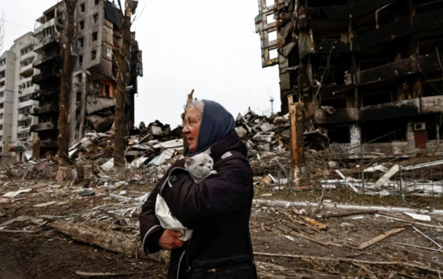 A woman carries her cat while standing in a street of bombed-out buildings