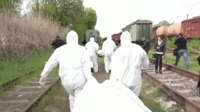 Several men in white clean suits carry white bodybags down the train tracks in this photograph