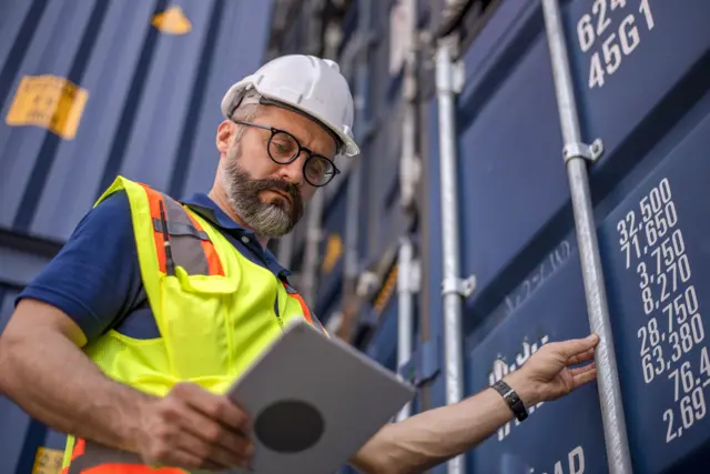 Man looks at tablet while holding handle of shipping container