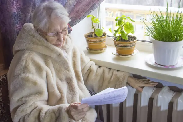 Older woman reads energy bill while sitting next to radiator in thick coat